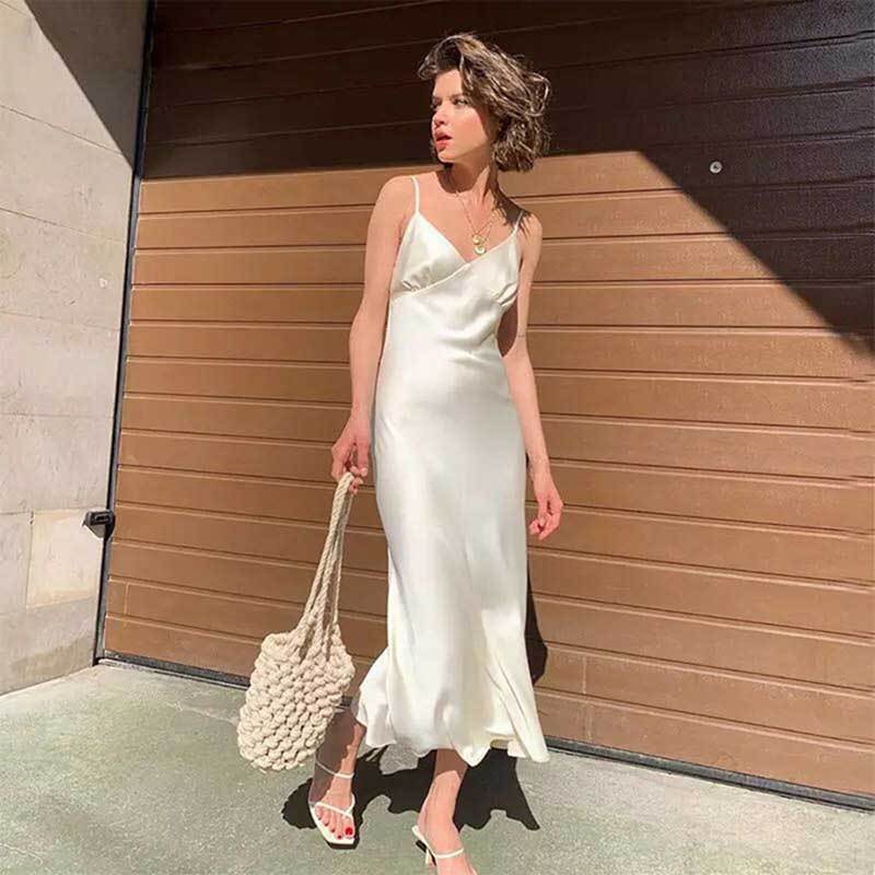 Woman wearing a white sleeveless long sling dress with a large swing skirt holding a woven bag outdoors