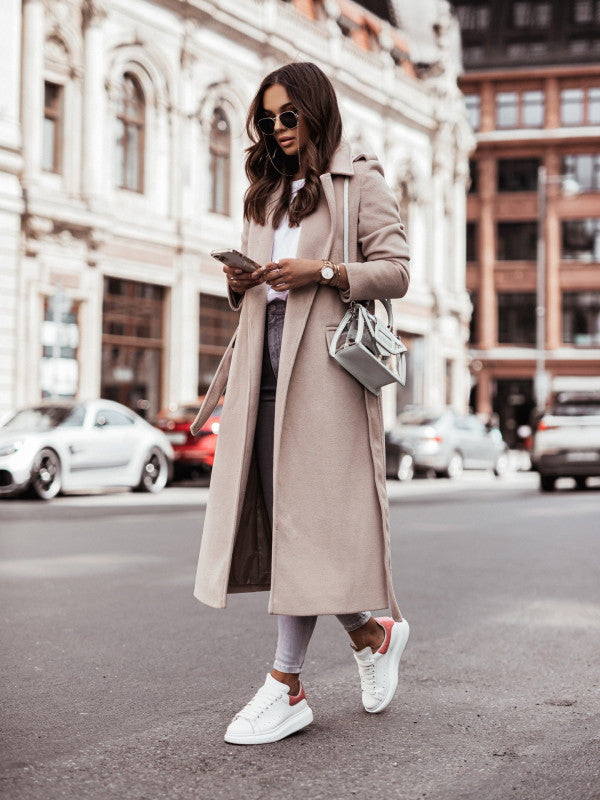 Women's long sleeve wool coat with waist tie, styled with sneakers and a handbag.